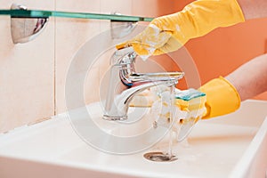 woman doing chores in bathroom at home, cleaning surfaces sink and faucet with spray detergent suds sponge.