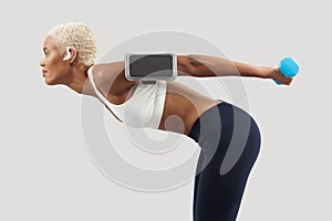 woman doing fitness workout, lifts dumbbells while listening music through earphones of mobile phone on arm band. Latin American