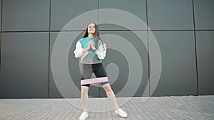 Woman doing fitness workout outdoor with rubber resistance band. Sportswoman in sporty leggings and top makes squats
