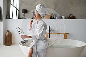 Woman doing makeup looking at mirror while sitting on bathtub