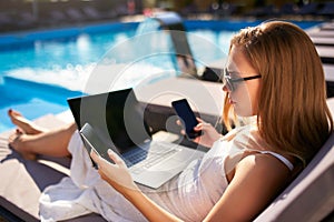 Woman doing remote multitasking work with multiple electronic internet devices on swimming pool beach bed. Freelancer