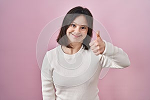 Woman with down syndrome standing over pink background doing happy thumbs up gesture with hand