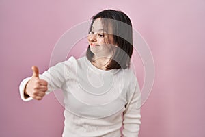 Woman with down syndrome standing over pink background looking proud, smiling doing thumbs up gesture to the side