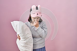 Woman with down syndrome wearing sleeping mask hugging pillow tired rubbing nose and eyes feeling fatigue and headache