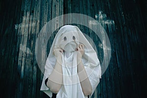 A woman draped in a white ghostly robe with holes for eyes, standing in front of a wooden church adorned with crosses on its doors