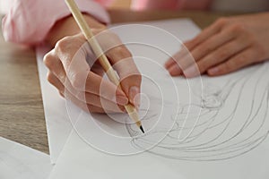 Woman drawing girl`s portrait with pencil on sheet of paper at wooden table, closeup