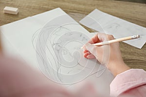 Woman drawing girl`s portrait with pencil on sheet of paper at table, closeup
