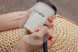 Woman drawing word donate on white cup. Hand holding cup of coffee take away. Donate logo on paper cup. Donation concept.