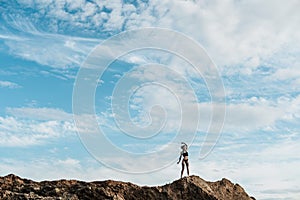 A woman with dreadlocks in underwear and tattooed stands on a sandy hill in the distance. Blue sky with clouds, copy