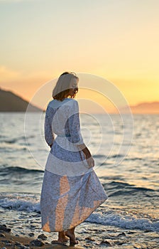Woman in dress standing at the sea beach. Nature and beauty concept. Girl silhouette at sunset