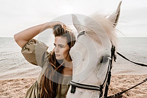 A woman in a dress stands next to a white horse on a beach, with the blue sky and sea in the background.