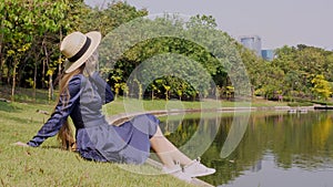 Woman in Dress and Straw Hat Sitting on Green Lawn Near Lake at Public City Park