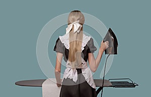 Woman dressed as a maid irons a towel on an ironing board. Back view. Blue background