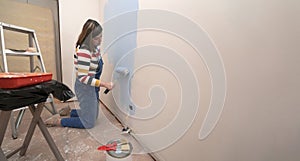 woman dressed in overalls and striped blouse, profile view, painting a white wall with a roller with blue paint, inside an empty