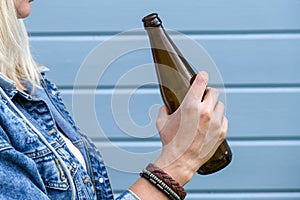 Woman dressed punk, infirmal holding a bottle of beer against blue wooden plank wall, abuse and female alcohol addiction concept