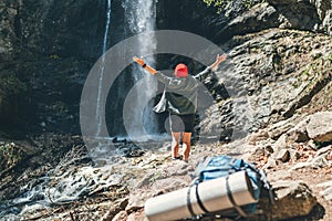Woman dressed in red hat and active trekking clothes near the mountain river waterfall rose arms up and enjoying the Nature.