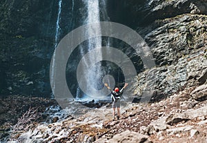 A woman dressed in a red hat and active trekking clothes standing near the mountain river waterfall rose arms up and enjoying the