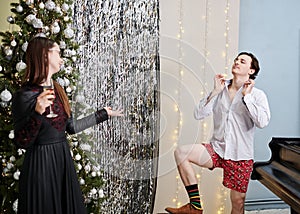 Woman,dressed up in fancy black dress with champagne glass surprised by man in shorts, shirt, socks showing off by Christmas tree