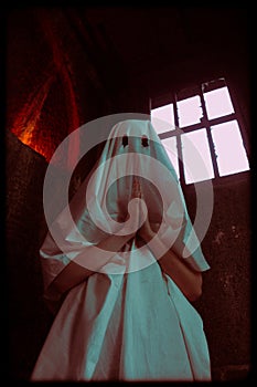 A woman dressed in a white ghostly attire with holes for eyes stands in a crypt, deep in prayer. The occultism, sects and religion