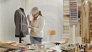 Woman dressmaker working with tailoring mannequin, sews the buttons to the jacket. With colorful clothes, fabrics and tools on the