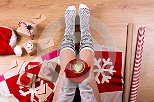 Woman drinking cappuccino coffee and sitting on the wooden floor. Close-up of female legs in warm socks with a deer with christmas