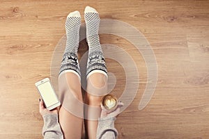 Woman drinking cappuccino coffee and sitting on the wooden floor. Close up of woman`s hands holding cup of coffee and using smart