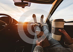 Woman drinking coffee paper cup inside car with feet warm socks on dashboard - Girl relaxing in auto trip reading travel book with