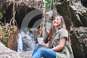 A woman drinking coffee while sitting on the rock in front of waterfall