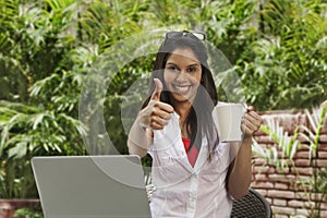 Woman drinking coffee, using a laptop and showing a thumbs up si