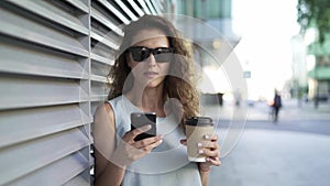 Woman drinking coffee and web surfing standing near gray wall in summer city
