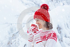 Woman drinking hot chocolate in Christmas morning in snowy garden. Girl in knitted Nordic sweater, hat and mittens holding cup