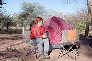 Woman drinking hot coffee mug while relaxing in camping site. Tent, chairs and camping gears. Outdoor activities in summer. Advent