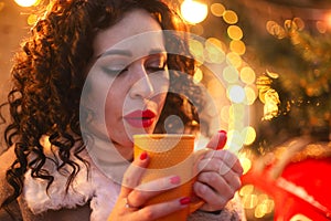 Woman drinking hot drink from mug while standing near glowing decorated Christmas tree outdoors