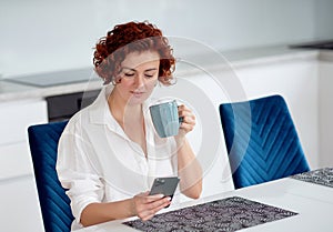 woman drinking tea and using smartphone in her kitchen while getting ready to go to work. messaging with friends while
