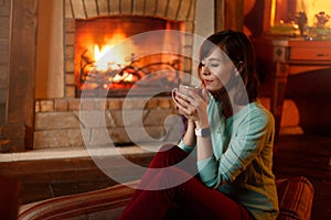 Woman is drinking tea and warming herself by the fireplace. Young caucasian female holds cup of coffee at home. Warm