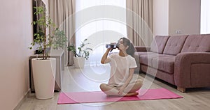 woman drinking water and sitting on yoga mat at home