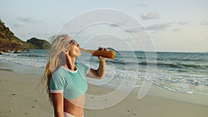 Woman drinking whiskey with orange juice from a bottle on beach, relax warm gold sunset. Woman walking in blue swimsuit
