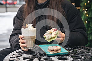 A woman drinks coffee with cream and eats Christmas macarons in a cafe outside.
