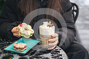 A woman drinks coffee with cream and eats Christmas macarons in a cafe outside.