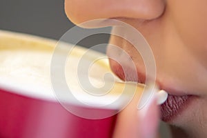 A woman drinks coffee with milk foam from a red paper Cup, selective focus, close-up.