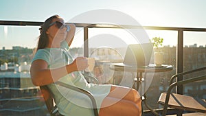 Woman drinks coffee and rests during a break on the balcony against the the setting sun. Remote work concept