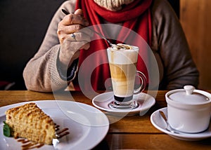 A woman drinks mokko coffee with cream of a transparent cup and a Napoleon cake in a white ceramic plate and white ceramic