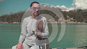 A woman drinks tea from a thermos in the sun, sitting on a wooden pier of the lake on a spring day, relaxing in nature.