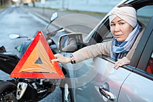 Woman a driver holding triangle sign is in her hand, sitting in car after road collision with motorcycle
