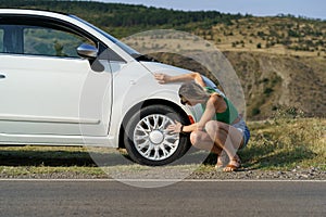 Woman driver parked white car on roadside of highway to check wheel tire on sunny summer day