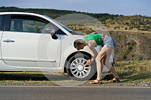 Woman driver parked white car on roadside of highway to check wheel tire on sunny summer day