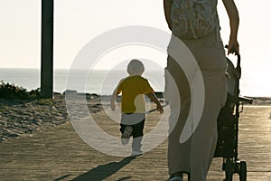 A woman drives a baby stroller and her little son runs forward to the ocean