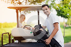 A woman is driving a golf car. A man in the foreground stands next to the trunk and pulls out a golf club