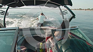 A woman is driving a motor boat, and in the background a guy is riding a wakeboard, and man is helping him