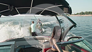 A woman is driving a motor boat, and in the background a guy is riding a wakeboard, and man is helping him